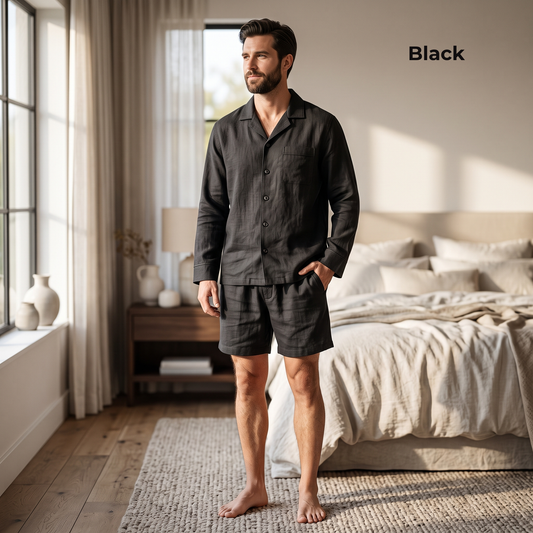 A man stands barefoot in a softly lit bedroom wearing a dark charcoal-black long-sleeve pajama shirt and matching shorts. The word 'Black' appears in the upper right corner, indicating the color option.