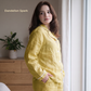 Woman wearing yellow pajamas standing in a room with a plant and wooden dresser.