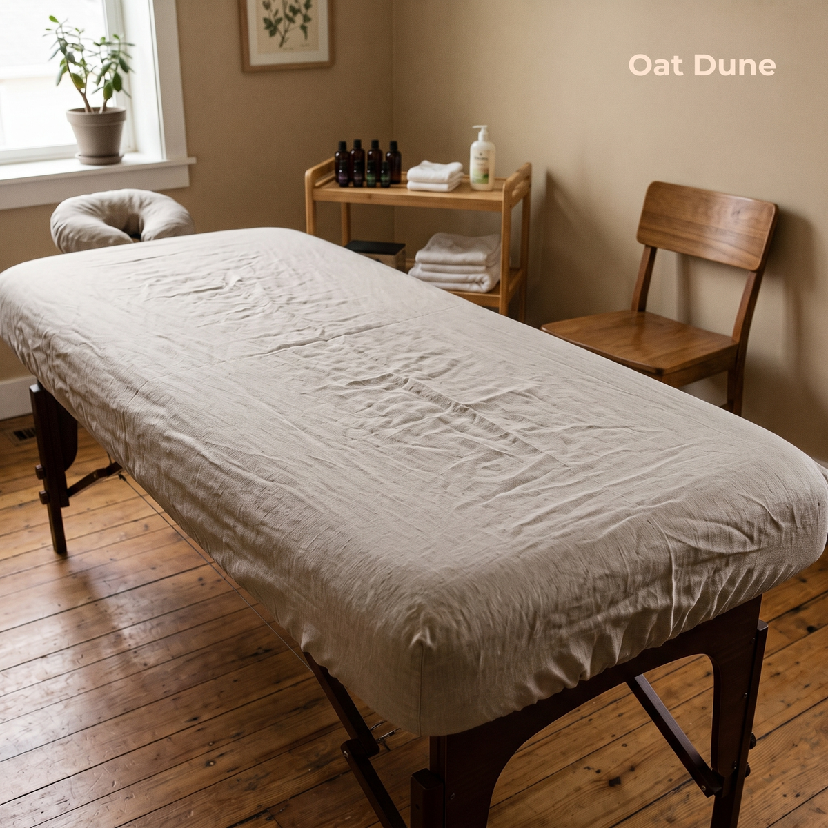 A massage table dressed with a light beige linen sheet, set in a warm-toned room with wooden floors, a wooden chair, and a small wooden side table holding towels and bottles.
