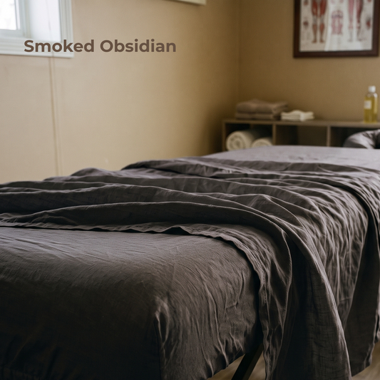 A bed dressed in dark charcoal gray bedding, shown in a softly lit bedroom with beige walls, a small shelf, and a framed picture in the background. The text 'Smoked Obsidian' appears in the upper left.