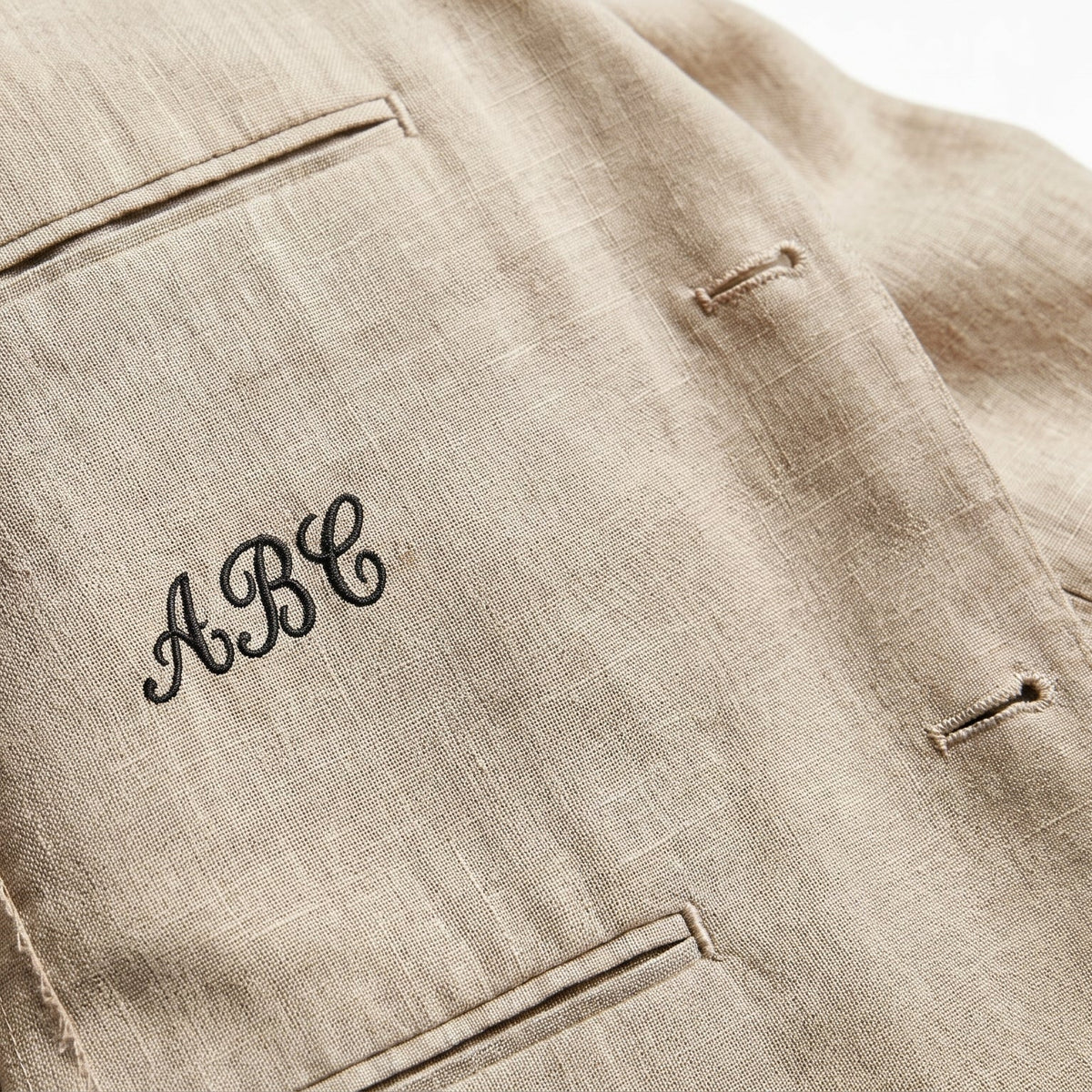 Close-up of a beige linen jacket with a black embroidered monogram 'ABC' on the pocket.