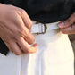 Close-up of a person's hands fastening a white belt with a brass buckle over a white shirt and dark pants by organolinen