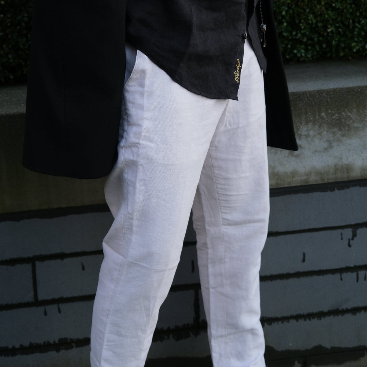 A person wearing white trousers and a black jacket, photographed outdoors on a paved walkway with greenery visible.