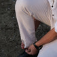 A close-up lifestyle shot of a person sitting on a textured concrete surface, tying the lace of a black leather dress shoe with white trousers and a white shirt.